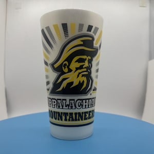 May include: A white tapered drinking glass featuring a black and yellow graphic of a bearded mountaineer wearing a hat. Behind the figure are radiating black and yellow sunburst lines. The text APPALACHIAN MOUNTAINEERS is printed in black.