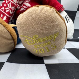 May include: Close-up of a plush toy's foot with the Disney Store 2015 logo embroidered in gold. The foot is tan with a brown trim, and the toy's red and white patterned sweater is visible.