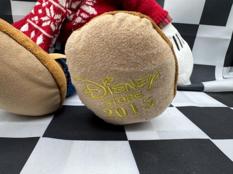 May include: Close-up of a plush toy's foot with the Disney Store 2015 logo embroidered in gold. The foot is tan with a brown trim, and the toy's red and white patterned sweater is visible.