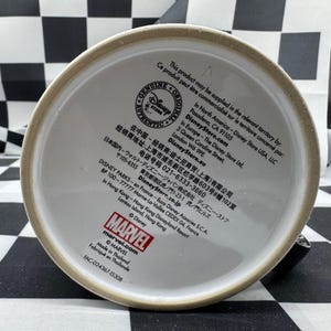 Puede incluir: La base de una taza de cer&aacute;mica blanca con un borde beige. La taza presenta texto, incluidos los logotipos de "Disney Store" y "Marvel", e informaci&oacute;n de fabricaci&oacute;n. La taza est&aacute; hecha en Tailandia.