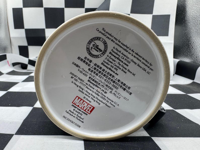 Puede incluir: La base de una taza de cer&aacute;mica blanca con un borde beige. La taza presenta texto, incluidos los logotipos de "Disney Store" y "Marvel", e informaci&oacute;n de fabricaci&oacute;n. La taza est&aacute; hecha en Tailandia.