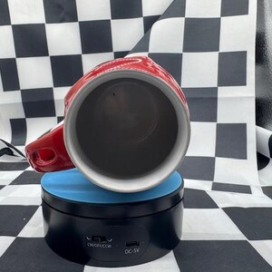 Puede incluir: Una taza de cer&aacute;mica roja y blanca con asa, colocada sobre un soporte giratorio azul y negro. El interior de la taza es oscuro y el asa est&aacute; en el lado derecho. El fondo presenta un patr&oacute;n de cuadros blancos y negros.