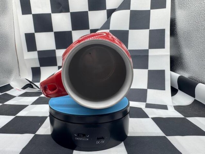 Puede incluir: Una taza de cer&aacute;mica roja y blanca con asa, colocada sobre un soporte giratorio azul y negro. El interior de la taza es oscuro y el asa est&aacute; en el lado derecho. El fondo presenta un patr&oacute;n de cuadros blancos y negros.