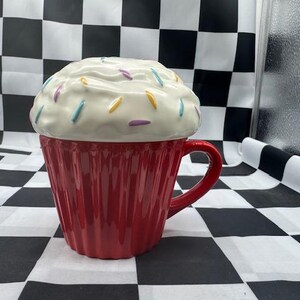 May include: A red ceramic cupcake-shaped mug with a white, textured lid decorated with colorful sprinkles. The mug has a handle and vertical ridged detailing, resembling a cupcake liner. The lid mimics frosting.