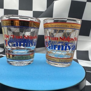 May include: Two clear shot glasses with gold and red accents. The glasses feature the text "The Fun Ships Carnival" in blue and red, with additional text in gold. The glasses are on a blue surface.