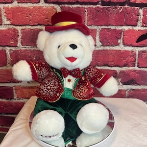 May include: A white teddy bear wearing a red top hat with a gold band, a red and gold jacket with snowflake designs, a green vest, and green pants. The bear has a red bow tie and is sitting on a mirrored surface.