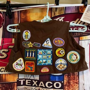 May include: A brown Girl Scout vest with multiple colorful patches. Patches include a penguin, soccer ball, and various event and achievement badges. The vest is hanging on a white hanger against a vintage-style background.