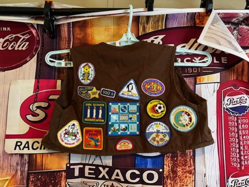 May include: A brown Girl Scout vest with multiple colorful patches. Patches include a penguin, soccer ball, and various event and achievement badges. The vest is hanging on a white hanger against a vintage-style background.