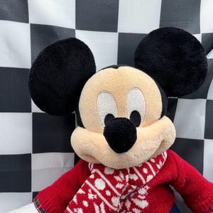May include: A plush Mickey Mouse doll with black ears, a tan face, and a black nose. The doll is wearing a red sweater with a white and red patterned scarf. The background is a black and white checkered pattern.