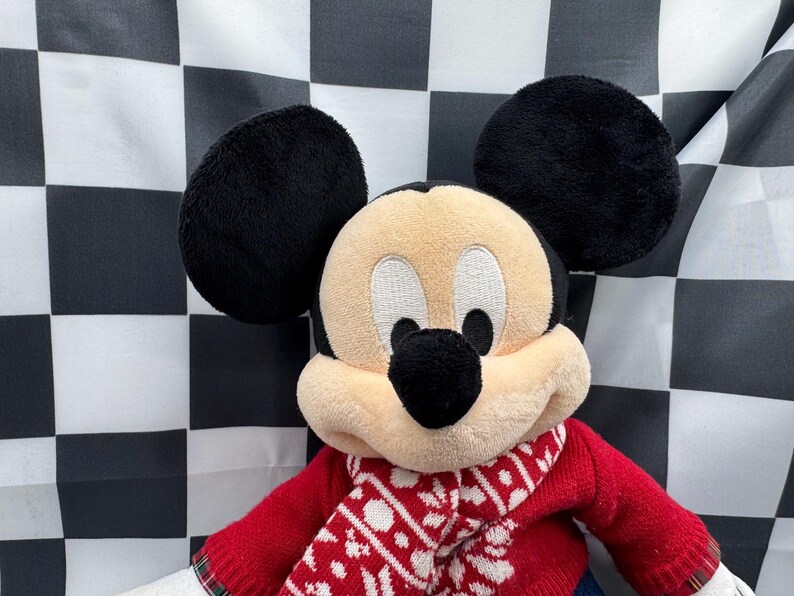 May include: A plush Mickey Mouse doll with black ears, a tan face, and a black nose. The doll is wearing a red sweater with a white and red patterned scarf. The background is a black and white checkered pattern.