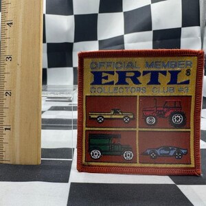 May include: A square patch with the text "OFFICIAL MEMBER ERTL COLLECTORS CLUB #3" in blue and yellow. The patch features illustrations of a yellow pickup truck, a red tractor, a green truck, and a blue sports car. A wooden ruler is next to the patch.