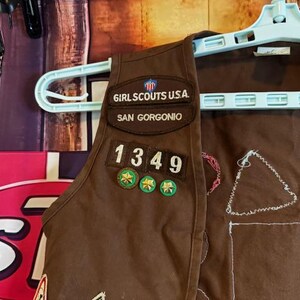 May include: A brown Girl Scouts vest with patches and the text "GIRL SCOUTS U.S.A. SAN GORGONIO 1349". The vest is hanging on a light blue hanger.