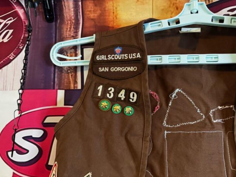 May include: A brown Girl Scouts vest with patches and the text "GIRL SCOUTS U.S.A. SAN GORGONIO 1349". The vest is hanging on a light blue hanger.