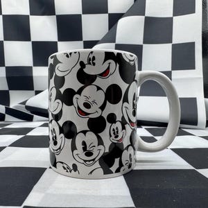 May include: White ceramic mug with a repeating pattern of Mickey Mouse faces in black and white. The mug has a white handle and is set against a black and white chequered background.