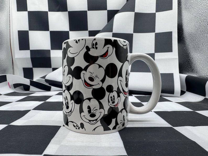May include: White ceramic mug with a repeating pattern of Mickey Mouse faces in black and white. The mug has a white handle and is set against a black and white chequered background.
