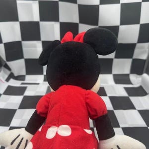 May include: A plush Minnie Mouse doll, viewed from the back, wearing a red dress with white polka dots and a red bow. The doll has black ears and arms, and white gloves. The background is a black and white checkered pattern.