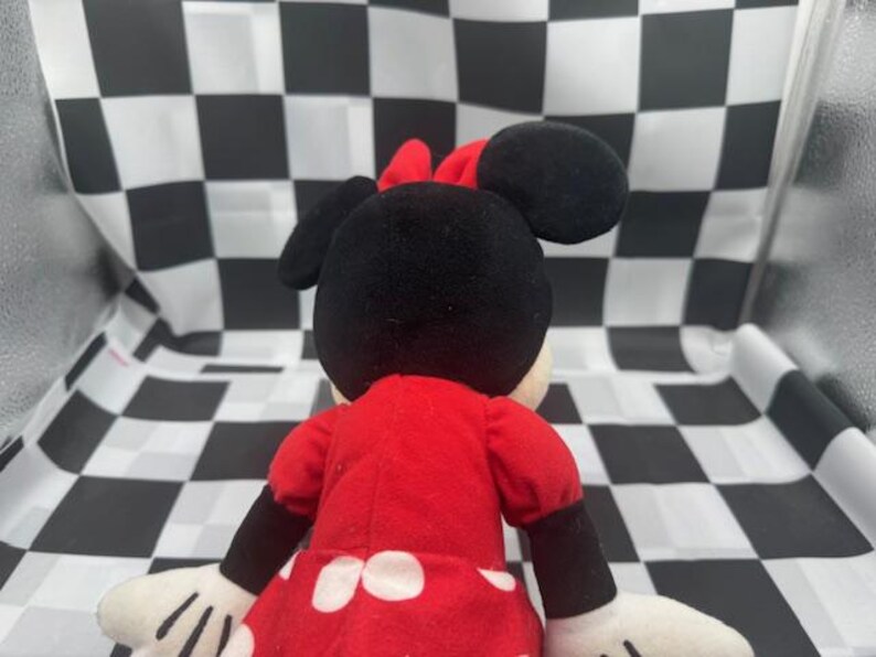 May include: A plush Minnie Mouse doll, viewed from the back, wearing a red dress with white polka dots and a red bow. The doll has black ears and arms, and white gloves. The background is a black and white checkered pattern.