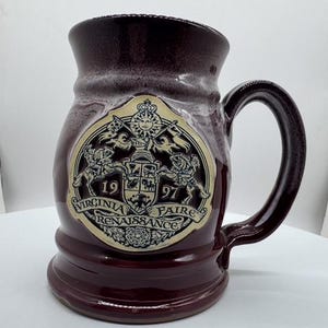 May include: A deep burgundy ceramic mug with a large, circular, cream-colored emblem on the front. The emblem features a sun, knights on horseback, and the text "1997 VIRGINIA RENAISSANCE FAIRE" in black lettering. The mug has a sturdy handle.