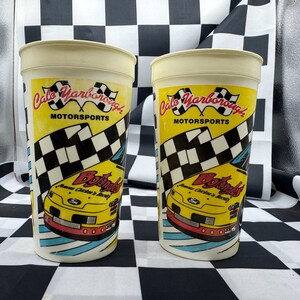 May include: Two vintage plastic cups with a racing theme. The cups are white with a yellow background and feature the text "Cale Yarborough Motorsports" and a checkered flag design. A yellow race car is also visible.