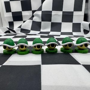 May include: Six small turtle figurines with green shells and white rims, yellow faces, and black eyes are arranged in a row. Each figurine sits on a green base with the word "PAWN" printed on it. The background features a black and white checkered pattern.