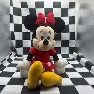 May include: A Minnie Mouse plush toy with a red dress with white polka dots, a red bow, and yellow shoes. The plush toy has black ears and a black nose. The background is a black and white checkered pattern.