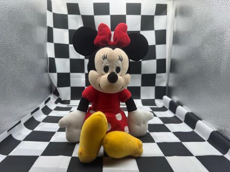 May include: A Minnie Mouse plush toy with a red dress with white polka dots, a red bow, and yellow shoes. The plush toy has black ears and a black nose. The background is a black and white checkered pattern.