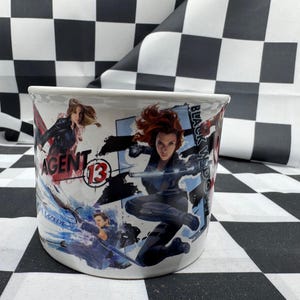 Puede incluir: Taza de cer&aacute;mica blanca con ilustraciones de acci&oacute;n de Black Widow y Agent 13. La taza tiene un dise&ntilde;o envolvente con las palabras "Black Widow" y "Agent 13" visibles. La taza es cil&iacute;ndrica y tiene un asa curva.