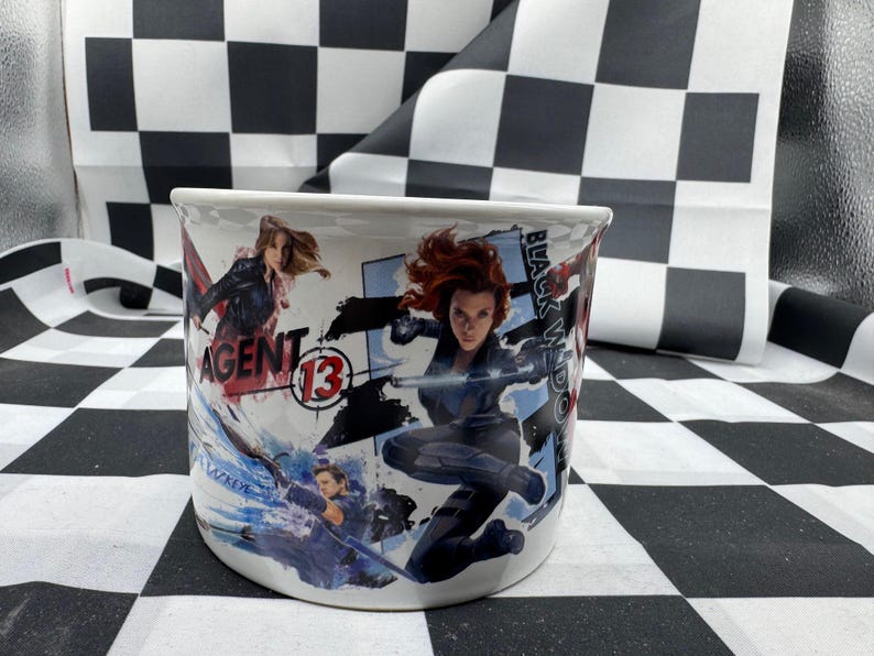 Puede incluir: Taza de cer&aacute;mica blanca con ilustraciones de acci&oacute;n de Black Widow y Agent 13. La taza tiene un dise&ntilde;o envolvente con las palabras "Black Widow" y "Agent 13" visibles. La taza es cil&iacute;ndrica y tiene un asa curva.