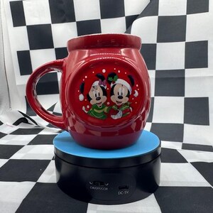 Puede incluir: Una taza de cer&aacute;mica roja con Mickey y Minnie Mouse con atuendos festivos. La taza tiene un asa grande y un dise&ntilde;o redondo con una imagen circular de los personajes. La taza est&aacute; sobre un soporte giratorio azul y negro.