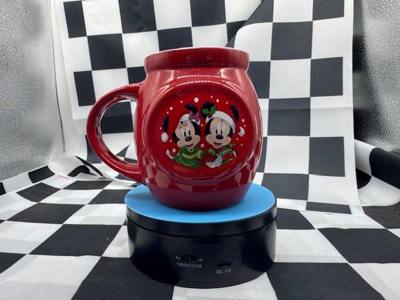 Puede incluir: Una taza de cer&aacute;mica roja con Mickey y Minnie Mouse con atuendos festivos. La taza tiene un asa grande y un dise&ntilde;o redondo con una imagen circular de los personajes. La taza est&aacute; sobre un soporte giratorio azul y negro.