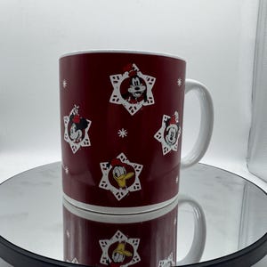 Pu&ograve; includere: Una tazza in ceramica bordeaux con manico e bordo bianchi. La tazza presenta immagini di Topolino, Minnie e Paperino in disegni a forma di fiocco di neve. La tazza &egrave; appoggiata su una superficie riflettente.