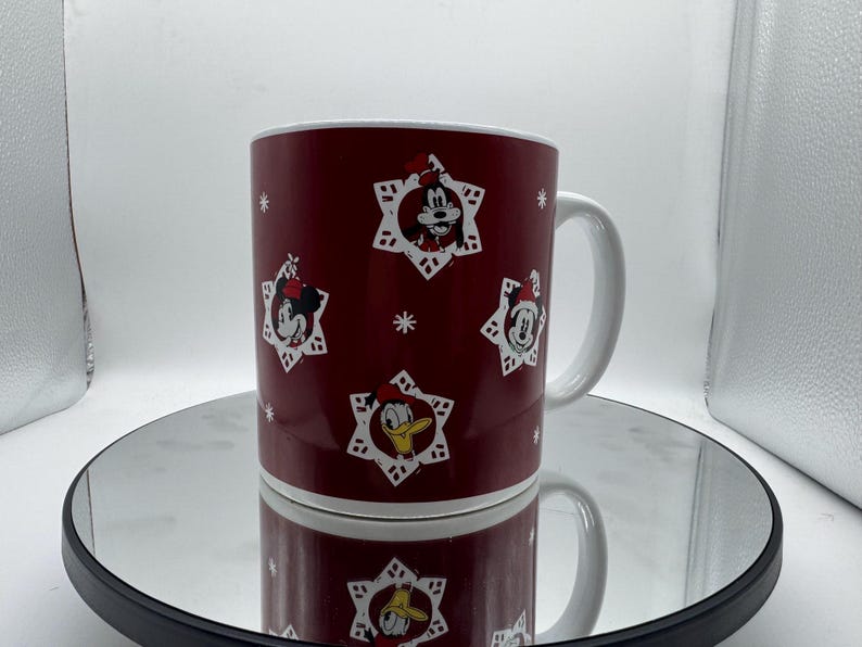 Pu&ograve; includere: Una tazza in ceramica bordeaux con manico e bordo bianchi. La tazza presenta immagini di Topolino, Minnie e Paperino in disegni a forma di fiocco di neve. La tazza &egrave; appoggiata su una superficie riflettente.