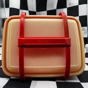 May include: A vintage Tupperware container in a rectangular shape, with a light yellow base and a red handle and strap closure. The Tupperware logo is visible on the red strap. The container is designed for food storage.