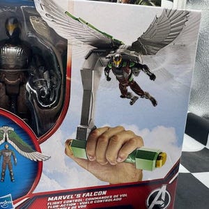 May include: Marvel's Falcon action figure in packaging. The figure is in a brown, green, and silver suit with wings. The packaging includes the text "Marvel's Falcon Flight Control". The figure is part of the Avengers series.