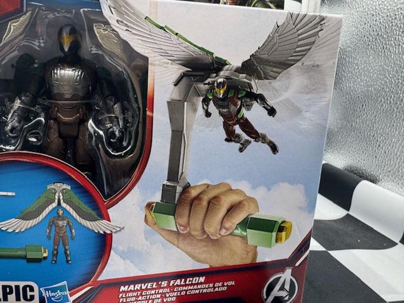 May include: Marvel's Falcon action figure in packaging. The figure is in a brown, green, and silver suit with wings. The packaging includes the text "Marvel's Falcon Flight Control". The figure is part of the Avengers series.