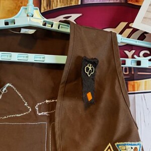 May include: A brown Girl Scout vest hanging on a light blue hanger. The vest has embroidered patches and a black and gold pin. The background includes a wooden wall and vintage signs.