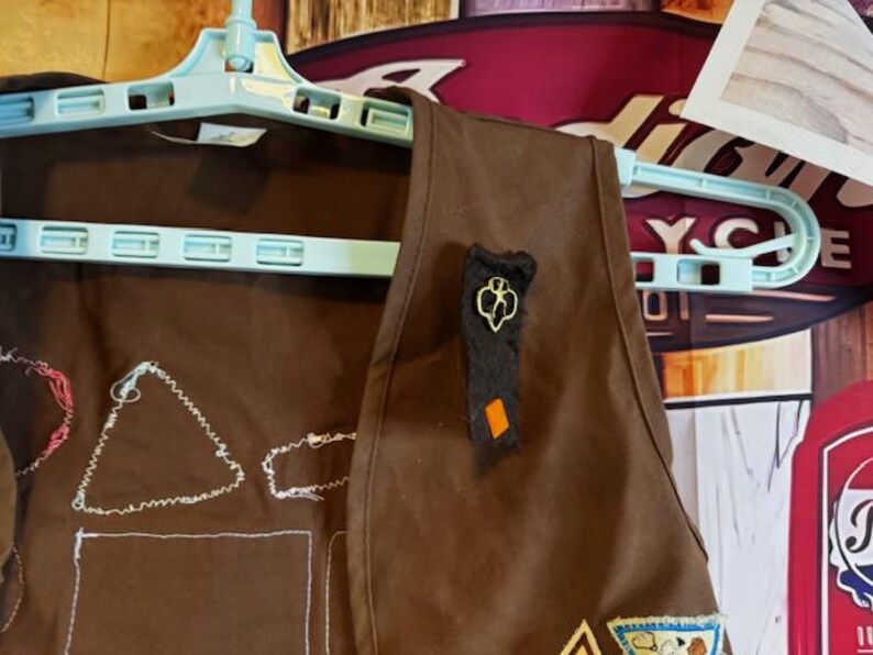 May include: A brown Girl Scout vest hanging on a light blue hanger. The vest has embroidered patches and a black and gold pin. The background includes a wooden wall and vintage signs.