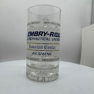 May include: A clear glass beer mug featuring the blue text EMBRY-RIDDLE AERONAUTICAL UNIVERSITY, the words Houston Center in a black serif font, and the word ALUMNI in blue, with a textured base and repeating university name text.