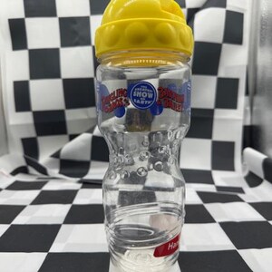 Peut inclure: Bouteille d'eau en plastique transparent avec un couvercle jaune. La bouteille a une forme incurvée et porte l'inscription "Ringling Bros. and Barnum & Bailey" en bleu et rouge. La bouteille est vide et porte une étiquette rouge en bas.