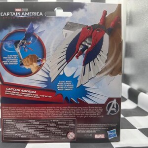Pode incluir: Uma embalagem de figura de a&ccedil;&atilde;o do Capit&atilde;o Am&eacute;rica da Marvel Studios. A caixa apresenta imagens do Capit&atilde;o Am&eacute;rica em voo, com asas vermelhas, brancas e azuis. O texto na caixa inclui "Captain America" e "Modo de Ataque!"