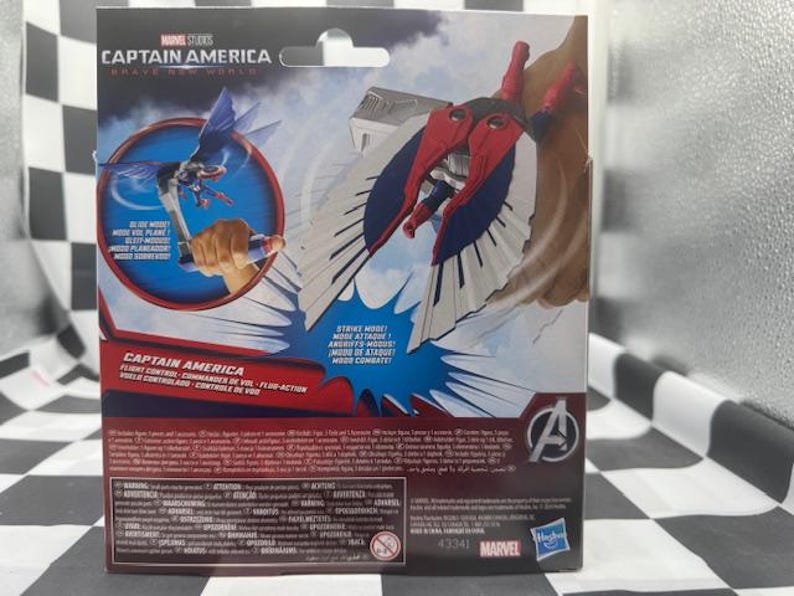 Pode incluir: Uma embalagem de figura de a&ccedil;&atilde;o do Capit&atilde;o Am&eacute;rica da Marvel Studios. A caixa apresenta imagens do Capit&atilde;o Am&eacute;rica em voo, com asas vermelhas, brancas e azuis. O texto na caixa inclui "Captain America" e "Modo de Ataque!"