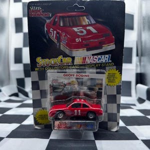 May include: A red die-cast NASCAR stock car model, number 11, in a clear plastic case, with a collector's card and display stand. The packaging features a larger image of a red race car, number 51, and the text "Racing Champions".