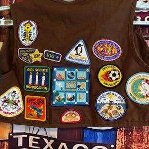 May include: A brown Girl Scout vest adorned with various colorful patches. Patches include a bird, penguin, soccer ball, and text that reads "Girl Scouts," "Early Bird 2000," and "Holiday Harmony 2000." The vest is hanging on a white hanger.