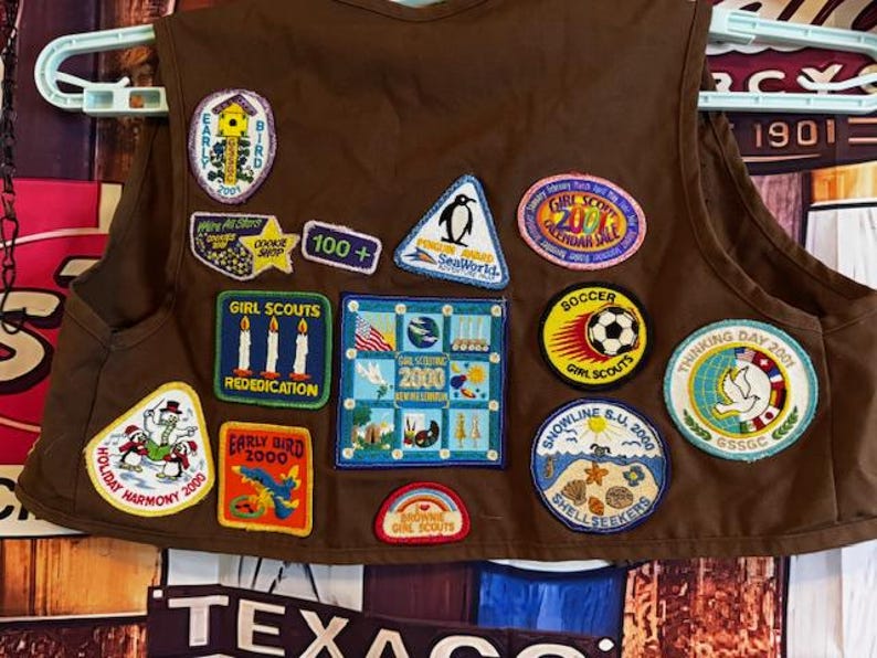 May include: A brown Girl Scout vest adorned with various colorful patches. Patches include a bird, penguin, soccer ball, and text that reads "Girl Scouts," "Early Bird 2000," and "Holiday Harmony 2000." The vest is hanging on a white hanger.