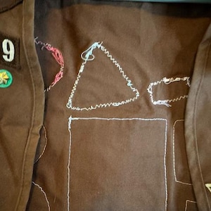 May include: Brown fabric with white stitched outlines of shapes, including a triangle and a rectangle. Patches with the number 349 and two green and gold pins are visible. The item appears to be a vest or similar garment.