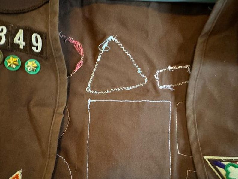 May include: Brown fabric with white stitched outlines of shapes, including a triangle and a rectangle. Patches with the number 349 and two green and gold pins are visible. The item appears to be a vest or similar garment.
