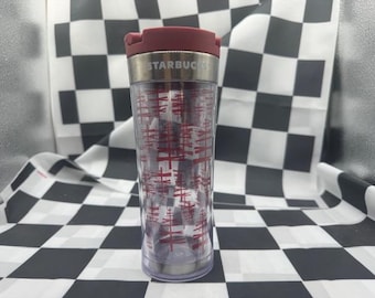 May include: A clear Starbucks travel tumbler with a maroon lid and a silver band. The tumbler features a red tree-like pattern. The Starbucks logo is visible on the silver band. The tumbler is on a black and white checkered surface.