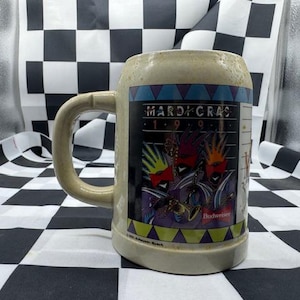May include: Tan ceramic beer stein with a large handle. The stein features a colorful graphic with the text "Mardi Gras 1999" and Budweiser branding. The design includes masked figures and musical instruments.