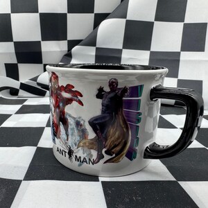 Puede incluir: Taza de cer&aacute;mica blanca con asa y borde negros. La taza presenta ilustraciones coloridas de personajes de c&oacute;mics, incluidos Iron Man y Vision. Las palabras "ANT MAN" est&aacute;n impresas en la taza.