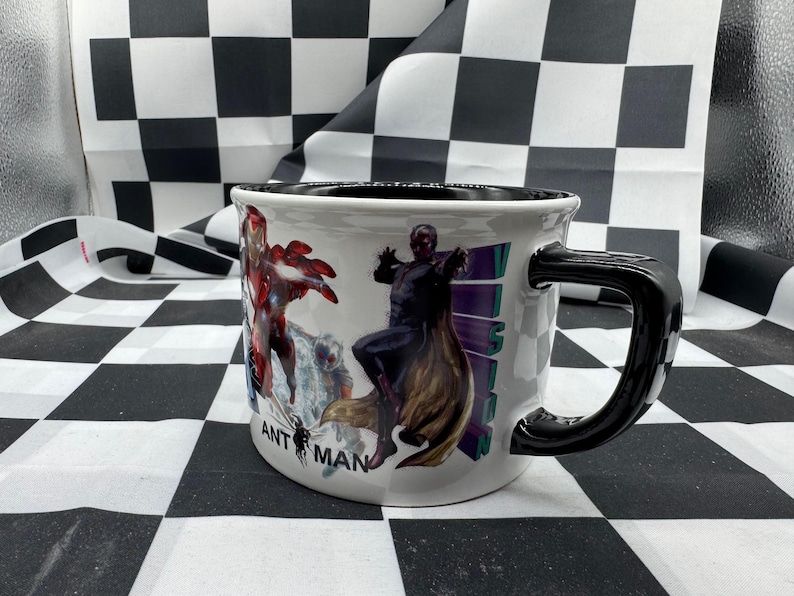 Puede incluir: Taza de cer&aacute;mica blanca con asa y borde negros. La taza presenta ilustraciones coloridas de personajes de c&oacute;mics, incluidos Iron Man y Vision. Las palabras "ANT MAN" est&aacute;n impresas en la taza.